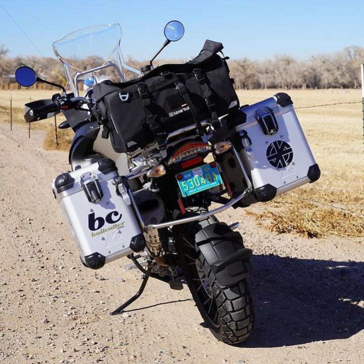 A black BMW adventure motorcycle loaded with travel gear, including a windshield, saddlebags, and side panniers, parked on a dirt road in a rural outdoor setting.